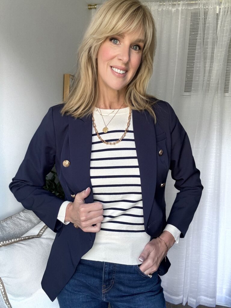 what to wear in paris this fall - Woman wearing a navy blazer over a striped sweater with gold jewelry and dark denim — classic French-inspired fall outfit.